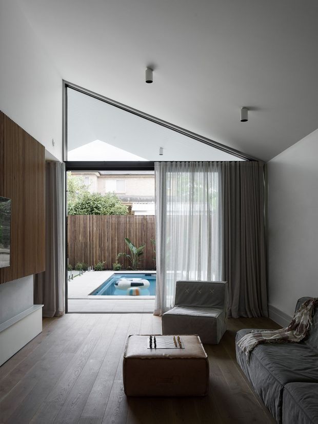 A contemporary living room with a low, sloping ceiling and dramatic floor-to-ceiling glazed opening leading to a private courtyard and small swimming pool. The room features pale timber flooring, white walls, dark timber vertical slat wall panelling, sheer and heavy grey curtains, a grey sofa, an upholstered armchair, and a central ottoman with a chess set. Minimal recessed and surface-mounted ceiling lights complete the restrained interior.