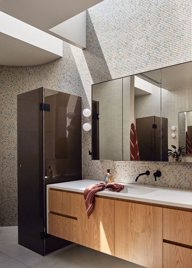 A contemporary bathroom with a long floating timber vanity, white benchtop, wall-mounted matte-black tapware, large mirror, and a dark glass shower enclosure. Small multicoloured mosaic tiles cover the walls, while strong natural light enters from a high opening or skylight, creating pronounced geometric shadows. The space has warm timber cabinetry contrasted with dark fixtures and glazing.