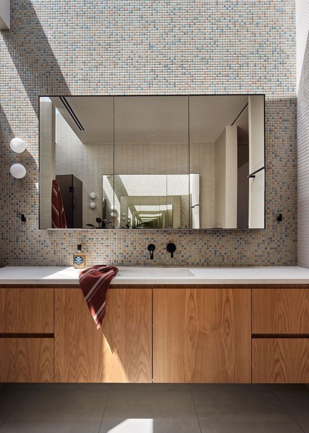 Contemporary bathroom vanity with a large wall-to-wall mirror, white benchtop, warm timber cabinetry, dark floor tiles, and a mosaic-tiled wall in muted blue, beige, grey, and terracotta tones. Recessed linear lighting and layered mirror reflections create depth, while a small towel and soap dispenser add lived-in detail.