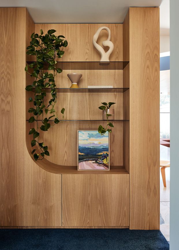 A close-up interior view of a custom light-oak timber built-in shelving and cabinetry unit. The shelving features dark glass shelves, a curved lower display shelf, integrated vertical storage panels, and carefully styled decorative objects including plants, artwork, a sculptural ornament, and an hourglass. A glimpse of an adjacent bright room is visible through the opening at right.