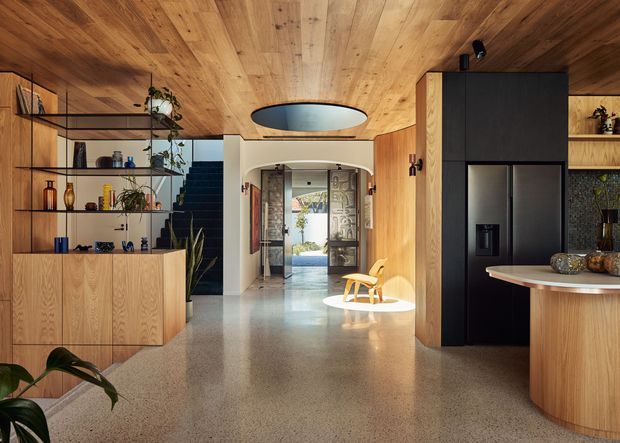 Warm contemporary open-plan kitchen and living area with extensive timber wall and ceiling lining, polished terrazzo-style flooring, integrated dark cabinetry and refrigerator, curved island benchtop, open shelving, and a central circular pendant skylight-like feature. A hallway and stairway are visible beyond the main space.