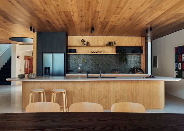 A contemporary open-plan kitchen dominated by warm timber panelling across the ceiling, island front, and rear cabinetry. The large island has a pale stone benchtop, integrated sink and tap, and timber stools. Dark built-in cabinetry, a textured blue-grey mosaic tile splashback, open timber shelving, and a tall dark refrigerator create contrast. A dining table is visible in the foreground, with a stairway and adjacent living area at left.
