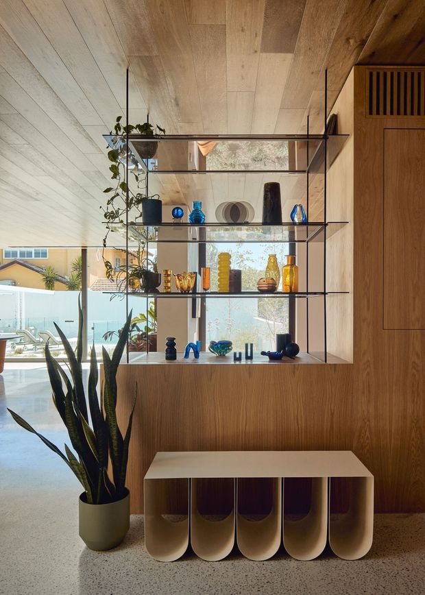 A warm, contemporary interior vignette featuring a full-height timber-lined built-in display wall with slim black metal shelving and glass shelves. The shelves hold colourful decorative vessels and ceramics, while a tall potted plant stands beside a sculptural cream storage bench. A large glazed opening provides views toward an outdoor pool area and neighbouring house. The floor is light speckled terrazzo, and integrated timber cabinetry includes a vented upper panel.
