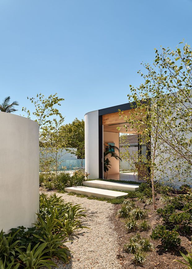 A landscaped outdoor approach to a contemporary pavilion-like entry, featuring curved smooth rendered walls, a dark flat roof edge, warm timber-lined ceiling, broad pale concrete steps, and a gravel garden path surrounded by drought-tolerant planting. A turquoise pool or water feature is partially visible beyond the entry, under a clear blue sky.