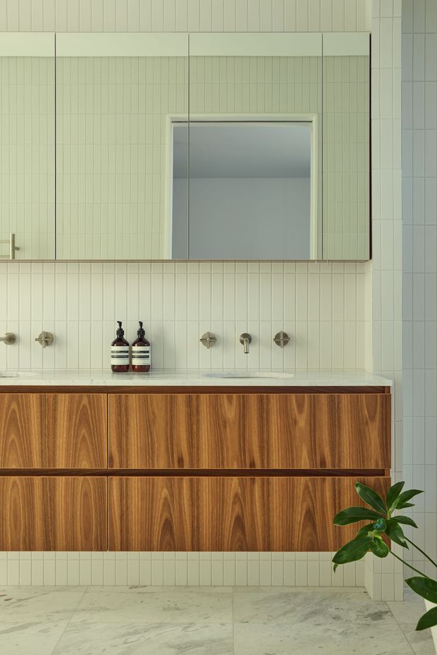 A close-up view of a refined bathroom vanity. The room features a wall-hung timber-grain floating cabinet with integrated basin and brushed metal wall-mounted tapware, set against pale vertical ribbed wall tiles. A large mirrored cabinet occupies the upper wall, reflecting the simple bathroom interior. The floor is finished in pale veined stone, and a potted plant adds greenery at the edge of the frame. The palette is muted, warm, and understated.
