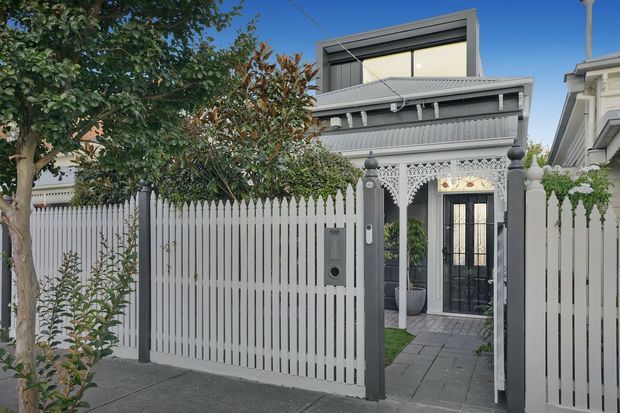 Street-facing view of a narrow urban residence combining a contemporary upper-level addition with a preserved ornate Victorian-style frontage. The property has a grey-and-white corrugated and rendered facade, a tall white picket fence, decorative wrought-iron-inspired entry detailing, and a paved path leading to glazed double entry doors.