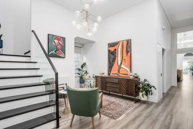 Bright, refined contemporary living and entry area with white walls, decorative ceiling moulding, pale timber flooring, and a sculptural globe pendant. A floating staircase with black metal handrails and glass balustrade occupies the left side, while a mid-century-inspired timber sideboard, green upholstered chair, patterned rug, plants, and colourful artwork create a warm, eclectic focal point. A hallway extends toward additional rooms in the background.