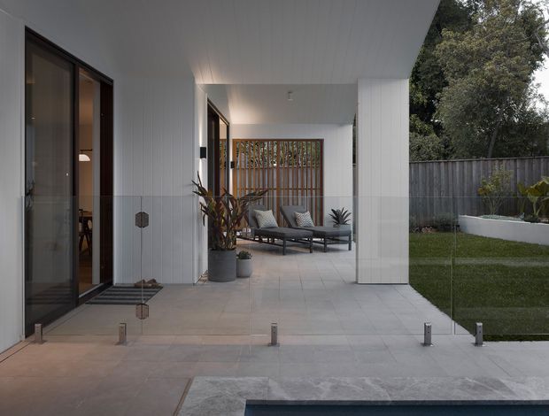 A contemporary covered outdoor patio and entry terrace viewed from beside a pool. The space features pale large-format tiled paving, white vertical wall cladding and a white paneled ceiling, dark-framed glazed doors, a timber slatted privacy screen, and a lounge seating area with potted plants. A fenced garden is visible beyond the covered area.