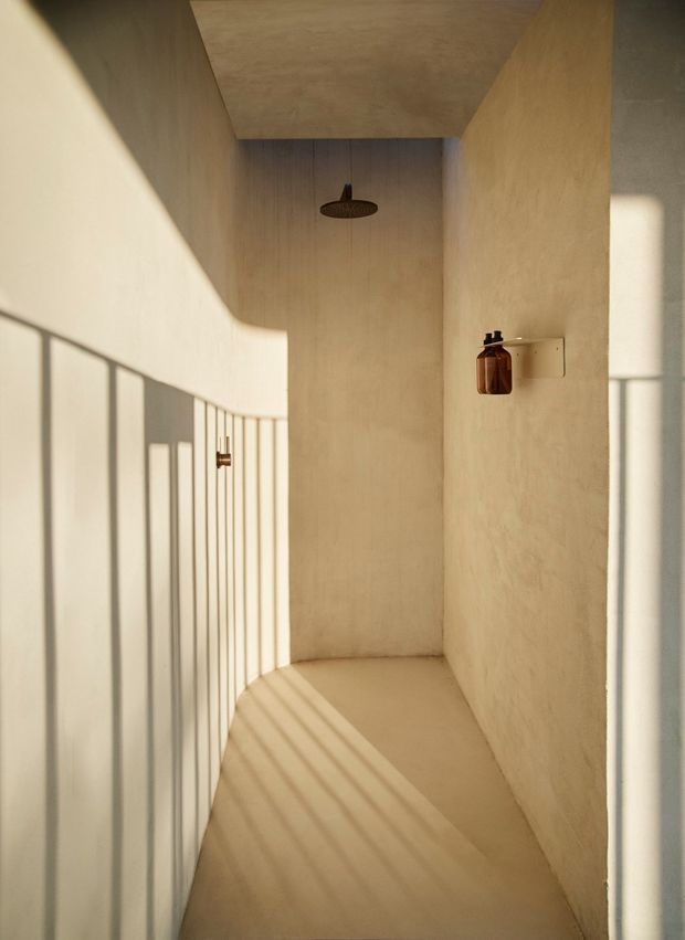 A narrow, minimalist shower room or wet-room corridor with continuous warm beige plaster or microcement-like surfaces on the walls, ceiling, and floor. A dark round rainfall showerhead is mounted overhead, with a small amber bottle on a wall shelf. Strong sunlight casts linear shadows from a slim vertical railing or screen across the left wall and floor, creating a calm, sculptural atmosphere.