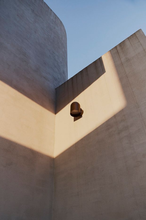 Minimal architectural view looking upward into a tall corner formed by smooth, warm-toned rendered or concrete walls. Strong angular sunlight creates a sharply defined geometric shadow across the walls, with a small dark cylindrical wall light mounted near the corner. A clear blue sky is visible above, emphasizing the sculptural, monolithic composition.