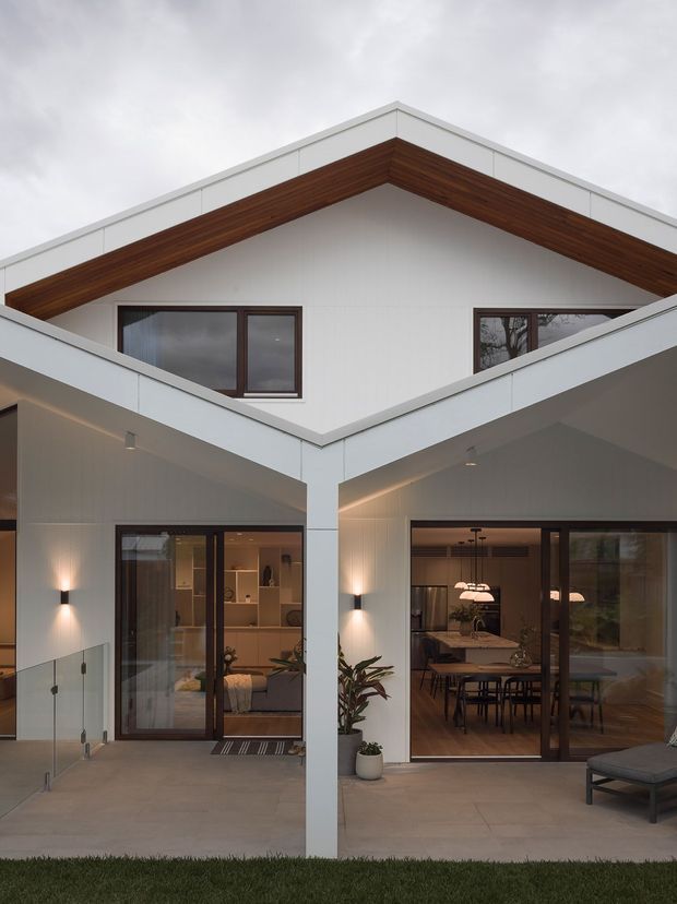 A contemporary two-storey residential facade viewed from an outdoor terrace or courtyard. The house has crisp white exterior wall and roof-edge cladding, strong angular gable forms, dark timber-lined eaves, and large dark-framed glazed openings. The lower level opens onto a broad concrete terrace with glass balustrading, wall sconces, potted plants, and views into a warmly furnished living and dining area. The upper gable contains dark-framed windows beneath a prominent timber soffit.