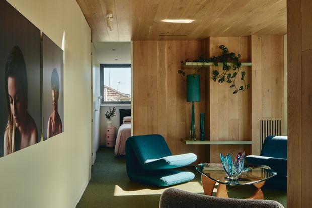 A warm, wood-lined living area viewed from a narrow hallway, featuring a teal lounge chair and matching seating around a glass coffee table with sculptural decor. Built-in timber shelving and cabinetry form a feature wall, with artwork along the hallway and a bedroom visible through the opening in the background. Natural light enters through a rear window, creating a refined mid-century-inspired interior.