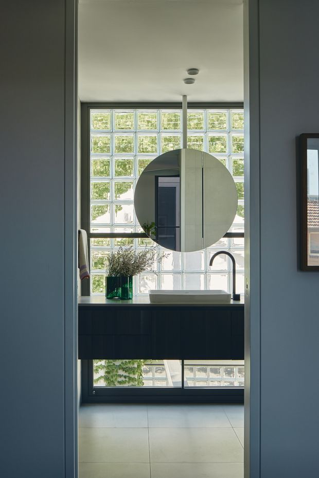 A compact, contemporary bathroom viewed through a doorway, featuring a wall-mounted dark vanity with a pale integrated basin, curved black tap, round wall mirror, and a large glass-block window providing diffused natural light. The room has light large-format floor tiles, dark blue-grey walls, and minimal recessed ceiling lights.