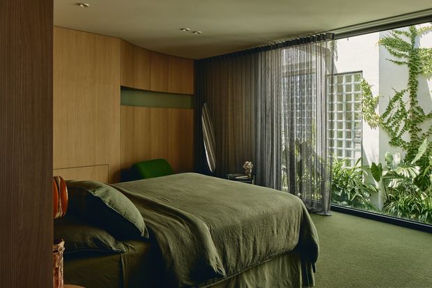 A moody, refined bedroom with a low-profile bed dressed in olive-green bedding, integrated warm timber wall panelling and cabinetry, and a dark green carpet. Full-height sheer and dark curtains frame expansive glazing overlooking a lush planted courtyard, bringing greenery into the room. A narrow recessed green feature niche and subtle built-in lighting complete the calm, cocooning interior.