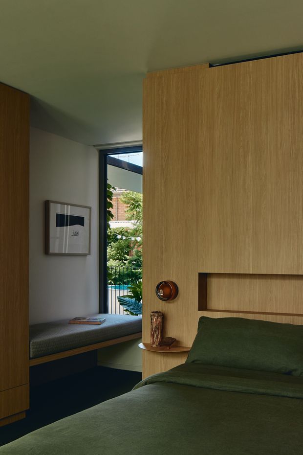 A compact, moody bedroom featuring a built-in timber bedhead and integrated bedside shelf, with warm vertical-grain wood cabinetry, dark green bedding, and a deep navy lower joinery band. A window seat occupies the adjacent alcove, and a narrow black-framed window provides views toward greenery and a pool area. The room has understated contemporary detailing and soft, muted natural tones.
