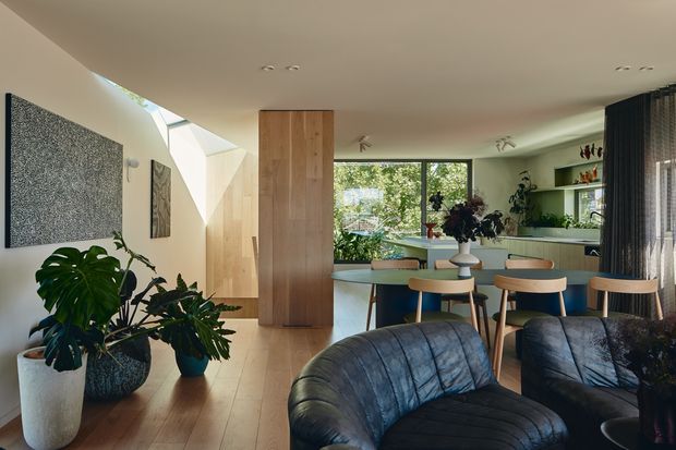 A bright, open-plan contemporary interior combining a dining area, kitchen, and living space. The room features warm timber flooring and a tall timber-clad partition or storage wall, a light neutral ceiling and walls, extensive glazing with garden views, a compact dining table with timber chairs, dark leather lounge chairs in the foreground, and abundant indoor plants. A textured wall artwork and sheer dark curtains add visual contrast.