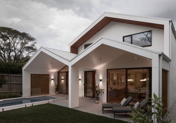 Exterior view of a contemporary multi-level home with a prominent gable roof, white rendered walls, dark timber soffit detailing, and extensive glazing. A covered outdoor entertaining area opens to a furnished living room, with a narrow swimming pool and landscaped lawn in the foreground. The overcast setting and warm interior lighting create a refined, inviting atmosphere.