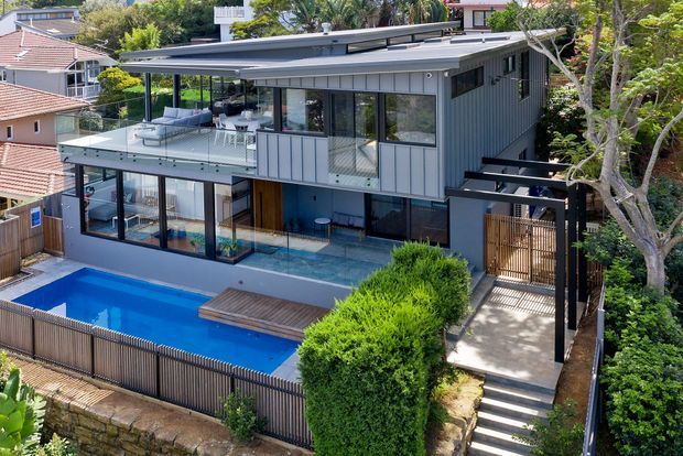 A multi-storey contemporary residential house on a sloping suburban site, viewed from above. The home has dark grey vertical cladding, extensive glazing, a broad low-pitched roof with a projecting eave, a glass-balustraded upper terrace, covered outdoor areas, and a bright blue swimming pool with a timber deck. Landscaped planting, retaining walls, and a stepped external path surround the property.