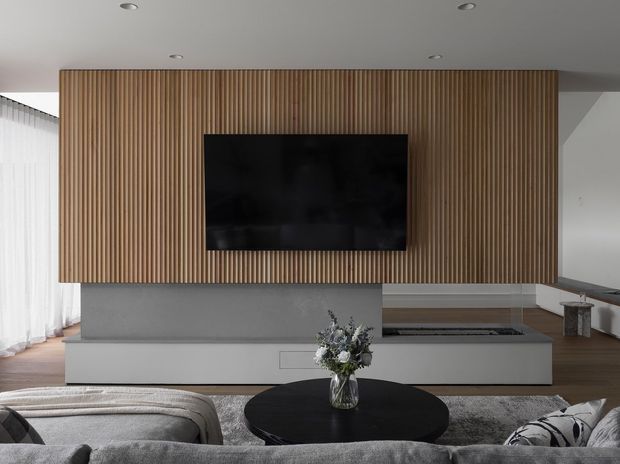 Contemporary living room viewed from behind a large sectional sofa, centred on a black round coffee table and a wall-mounted television. The TV is set into a full-height vertical timber-slat feature wall above a long minimalist grey built-in media cabinet and hearth-like joinery. Neutral grey furnishings, pale walls, recessed ceiling lights, timber flooring, sheer curtains, and a partial kitchen bench create a refined, minimalist interior.