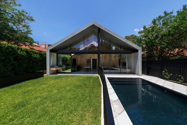 A contemporary pavilion-style backyard extension with a strong symmetrical gable roof and exposed concrete frame. Full-height glazing opens onto a manicured lawn and narrow swimming pool, while warm timber-lined interiors are visible through the glass. Established greenery, black fencing and neighbouring terracotta roofs surround the structure.