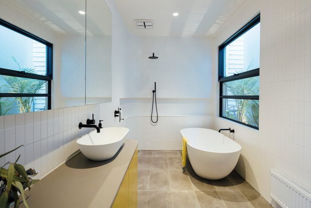 Bright contemporary bathroom with twin freestanding vessel basins on a long vanity, a freestanding bathtub, wall-mounted shower, white tiled walls, large black-framed windows, and a full-width mirror. The room has a clean, airy palette with warm neutral floor tiles and yellow vanity accents.