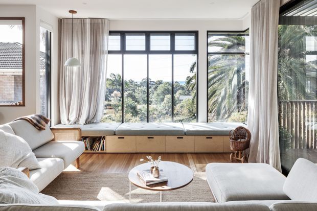 Bright, airy living room with large black-framed windows, a built-in timber window seat with drawers and shelving, pale upholstered seating, a round coffee table, timber flooring, and full-height sheer curtains. The room overlooks lush greenery and is styled in a calm, contemporary manner.