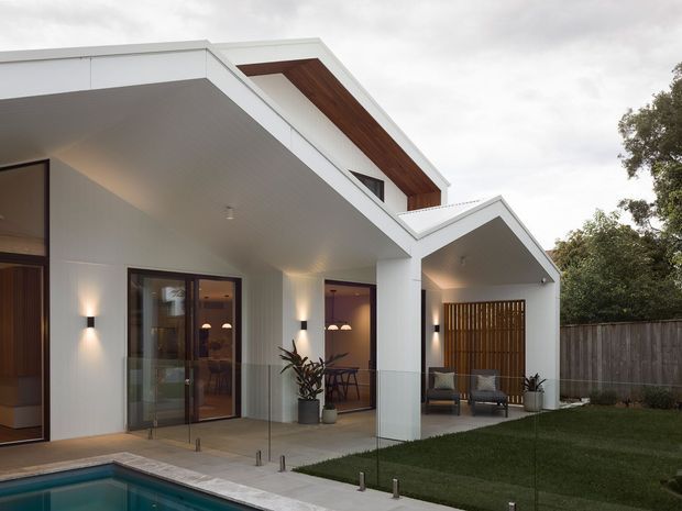 Contemporary two-storey residential exterior viewed from a landscaped backyard, featuring crisp white rendered walls, projecting flat roof forms, timber soffit detailing, dark-framed glazing, a covered outdoor seating area screened by vertical timber battens, and a swimming pool in the foreground.
