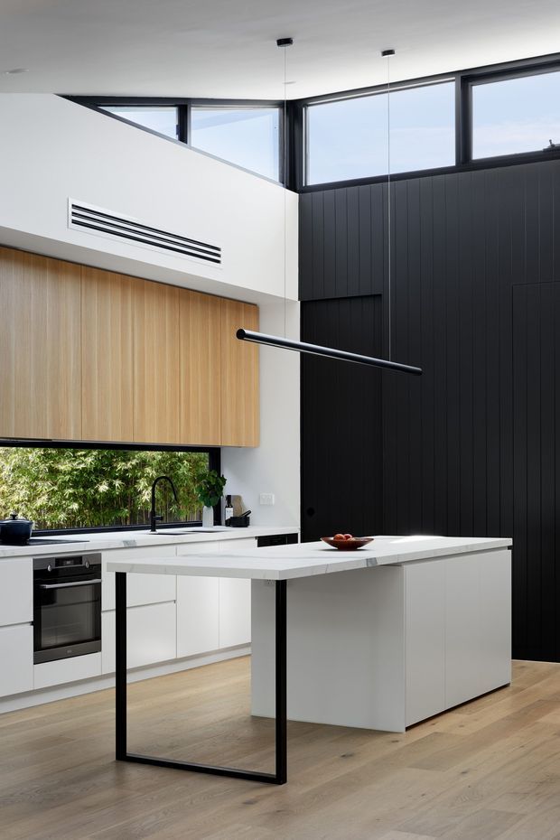 A contemporary kitchen with a large white island featuring a pale stone benchtop and black metal frame detail. White cabinetry and integrated appliances sit beneath light timber overhead cabinetry, while a dramatic black vertically panelled wall and black-framed clerestory windows add contrast. The room has light timber flooring, a black gooseneck tap, minimalist pendant lights, and abundant natural light.