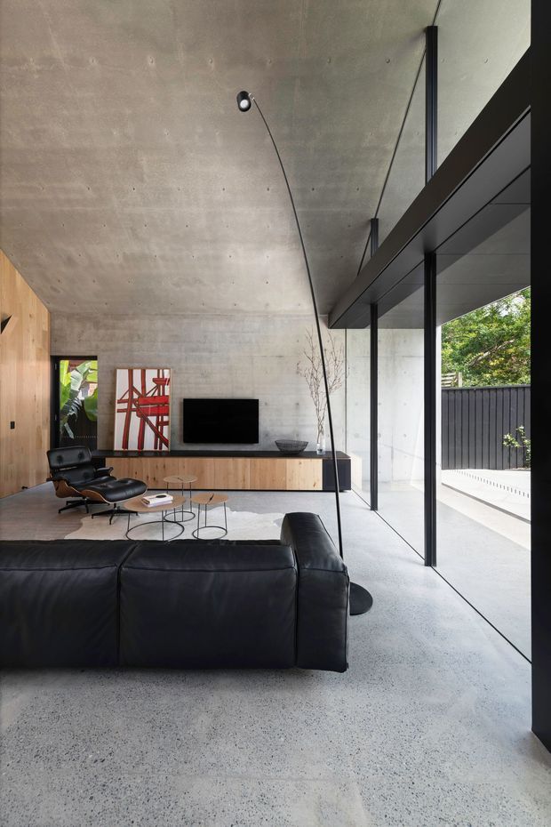 A refined modern living room with a large black leather sectional, low round coffee tables, a timber media console, wall-mounted television, and sculptural lounge chair. The space features an exposed concrete ceiling and walls, polished concrete or terrazzo-like flooring, extensive black-framed glazing opening toward an outdoor area, and minimalist integrated lighting. A red abstract artwork provides a small colour accent.