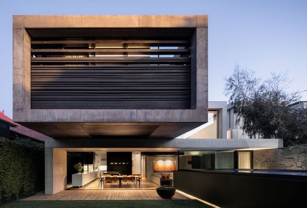 Exterior view of a contemporary multi-level residence with a strong cantilevered upper volume, exposed concrete framing, dark horizontal louvres, and extensive ground-floor glazing opening onto a timber deck. The lower level appears to contain an open-plan kitchen and dining area, with a dark boundary wall and a stone-clad side wall. The composition emphasizes bold geometric forms, concealed lighting, and indoor-outdoor connection.