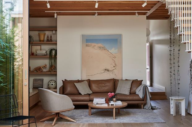 Warm, carefully styled living room with a brown leather sofa, sculptural upholstered lounge chair, round timber coffee table, and neutral rug. The room features extensive timber ceiling lining and flooring, built-in open shelving, indoor planting, full-height glazing, and a vertical slatted screen with trailing plants. A large desert-toned artwork provides a subtle focal point. Track lighting and natural light create a refined, relaxed atmosphere.