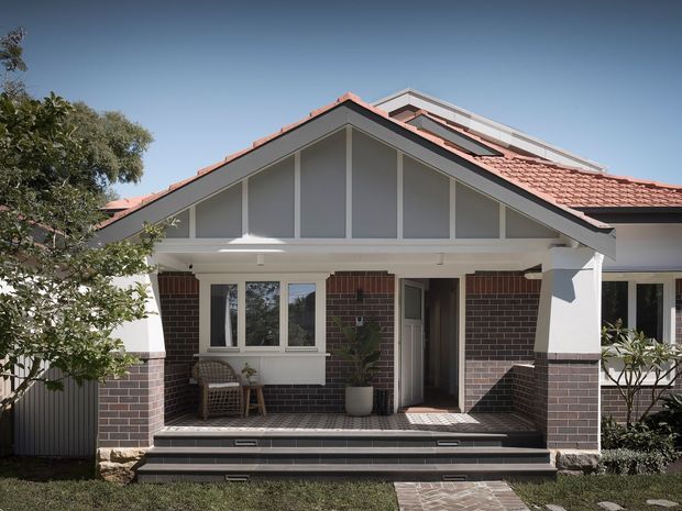 Front exterior view of a suburban brick home with a prominent gabled porch and symmetrical decorative timber detailing. The facade combines dark brick walls, white-painted trims and posts, a terracotta tiled pitched roof, and a raised tiled entry terrace with steps. A glazed upper gable and roof elements are visible, with limited landscaping around the entrance.