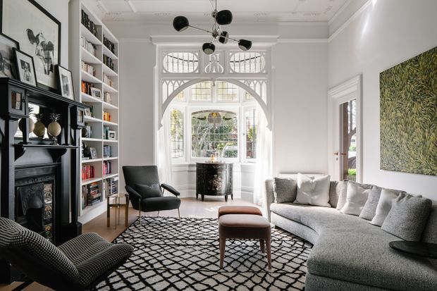 Bright, elegant living room in a period home, featuring a black traditional fireplace with mantel, full-height built-in bookshelves, ornate white ceiling cornices, decorative leadlight-style arched glazing, a large contemporary sectional sofa, patterned rug, sculptural black pendant light, and a botanical wall artwork. The space blends heritage architectural detailing with refined contemporary furnishings.