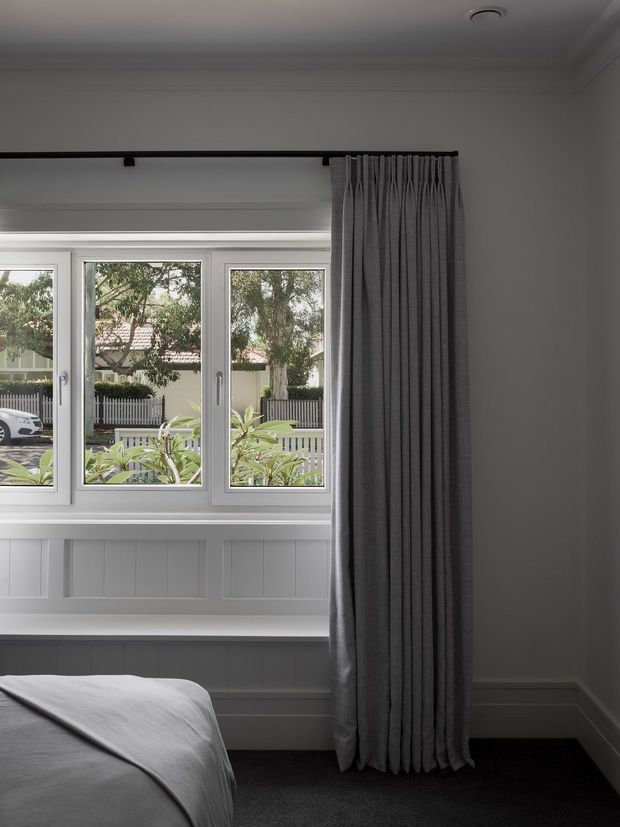 A softly lit bedroom corner with a low bed partially visible in the foreground, a large white-framed multi-pane window, built-in window seat with vertical wall panelling, and full-length dark grey pleated curtains on a black curtain rod. The room has ornate ceiling cornices, white walls and trim, and a dark carpet, creating a restrained traditional interior.