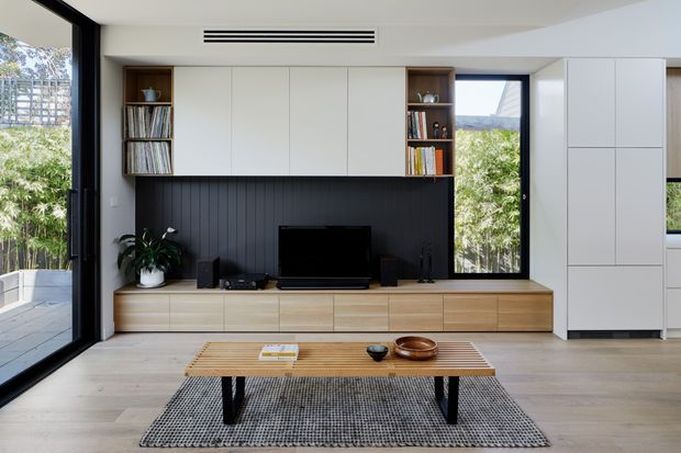 A bright, contemporary living room centered on a built-in entertainment wall. White overhead and side cabinetry surrounds open timber display niches, while a dark vertical-panel feature wall frames the television. A low timber coffee table, textured grey rug, pale timber flooring, indoor plant, and large black-framed glazed openings create a clean, warm, indoor-outdoor setting.