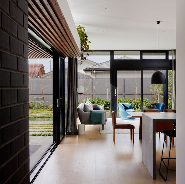 Contemporary open-plan living and dining area with full-height black-framed glazing opening to a landscaped courtyard. The room features pale timber flooring, a dark exposed brick wall, a timber slatted ceiling detail, a concrete kitchen island, and a mix of blue and neutral upholstered seating. A dining table and chairs occupy the foreground, while a lounge seating area sits beyond the glazing line.