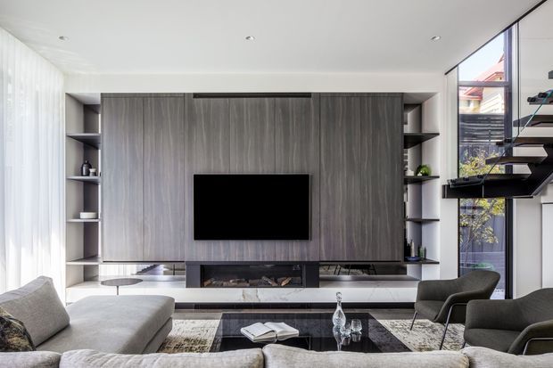 Contemporary living room with a large grey sectional sofa, dark lounge chairs, a black coffee table, and a full-height timber-look media wall with integrated shelving, television, and linear fireplace. Floor-to-ceiling glazing with sheer curtains brings in abundant daylight, while a floating open-riser stair is visible at the right.