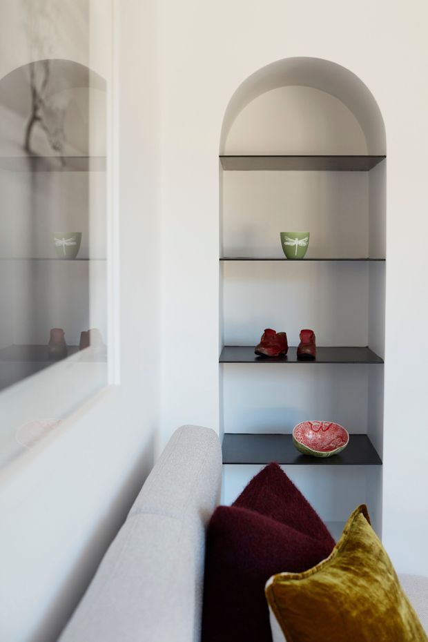 Interior detail of a refined living room showing a built-in recessed display shelving unit with a rounded arch top and dark shelves. Small decorative objects are arranged on the shelves, while the foreground includes the arm and cushions of a pale upholstered sofa. A large reflective glazed or mirrored surface appears on the left, echoing the shelving and room details. The space has a restrained neutral palette with burgundy and mustard accent cushions.