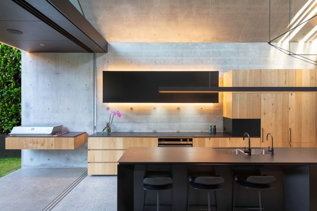 A refined contemporary kitchen with a large dark island and three stools, pale timber cabinetry, a black benchtop, and a matching black wall-mounted feature unit. Exposed concrete walls and ceiling create an industrial architectural backdrop, while full-height glazing and a garden view bring in natural light. A compact utility or appliance zone is visible at the left.