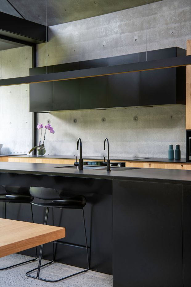 A sophisticated modern kitchen with a large black island, integrated dark cabinetry, and two slim black gooseneck taps over the sink. The space features exposed concrete walls and ceiling, a warm timber benchtop or rear work surface, black pendant lighting with a subtle brass accent, and black bar stools. A timber dining table is visible in the foreground, with orchids and small decorative bottles adding limited colour.