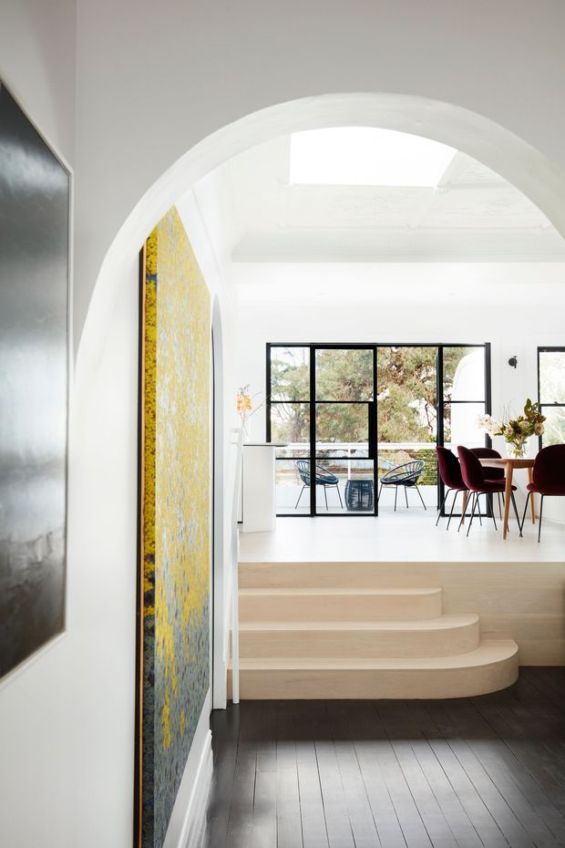A bright interior view through a large white arched opening into an elevated dining and sitting area. Curved pale timber steps lead up from a dark timber-floored space, with black-framed glazing, a skylight, white walls, a dining table with burgundy upholstered chairs, and a small balcony visible beyond. Large colourful artwork lines the left wall.