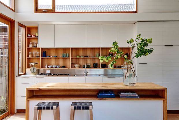 Bright contemporary kitchen with extensive white flat-panel cabinetry, warm timber open shelving and a substantial timber island bench. A stainless-steel cooktop, sink and integrated appliances line the rear wall, with two stools at the island. Timber-framed windows bring in abundant natural light, and a white horizontal-panel wall adds texture.