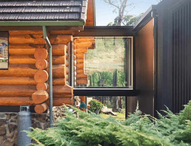 Exterior architectural detail showing a rustic log cabin joined to a dark contemporary extension. The cabin has rounded timber logs, a stone base, green-painted gutters and a shingle roof, while the extension features dark vertical cladding and large black-framed glazing overlooking a wooded setting. Ferns and planting occupy the foreground.