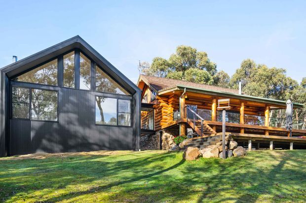 Exterior view of a rustic timber log cabin beside a modern dark corrugated-metal extension. The cabin has a pitched roof, exposed log walls, timber deck and balcony railings, while the adjacent extension features a prominent gabled glazed facade and dark vertical ribbed cladding. The buildings sit in a leafy landscaped setting.