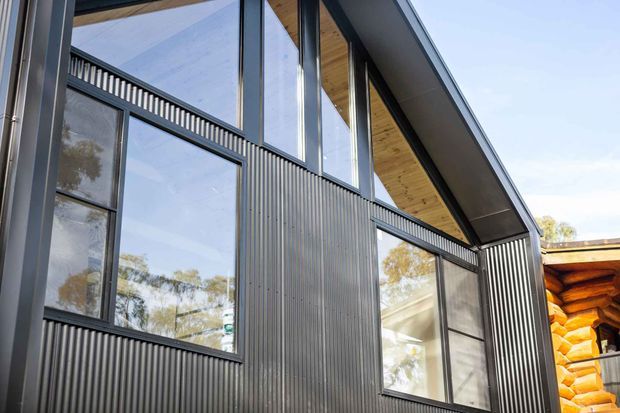 Exterior close-up of a contemporary gable-roofed building with dark vertically corrugated metal cladding, black-framed glazing, and a warm timber-lined soffit. A contrasting log construction is visible at the adjoining structure on the right, while trees are reflected in the windows.