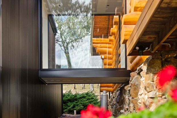 Exterior architectural detail of a rustic log-and-stone building, showing a large dark-framed glazed connection between a vertically ribbed dark wall and a timber log structure. A projecting glass volume or bridge sits above a rough natural-stone wall, with exposed timber rafters and beams visible beneath the roof. Landscaping and trees appear around the building, with blurred red flowers in the foreground.