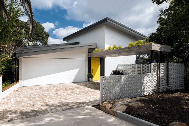 Contemporary two-storey residential exterior with a clean white rendered facade, broad low-pitched roof planes, an integrated double garage, and a bright yellow entry door. A paved driveway leads to the home, while a timber-toned pergola and patterned breeze-block privacy screen define the front garden and entry approach. Mature tropical and subtropical planting surrounds the property.