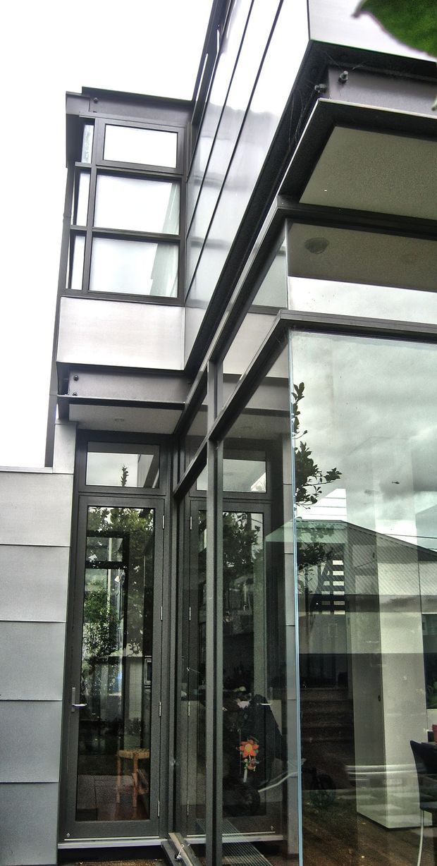 Vertical exterior view of a contemporary multi-level residential facade. Dark-framed glazing, a tall glazed entry door, projecting upper-level window bays, and reflective metal cladding define the composition. A glazed corner reveals part of the interior, while deep boxed eaves and balcony-like projections create strong horizontal and vertical lines.