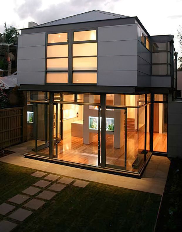 Exterior view of a contemporary two-storey urban residence at dusk. The upper level has a dark hipped roof, large warm-lit rectangular windows, and pale horizontal-panel cladding. The ground floor is largely enclosed by floor-to-ceiling glazing with dark metal frames, revealing an open-plan interior with timber flooring, a pale kitchen island, and a staircase. A paved path and small lawn occupy the foreground.