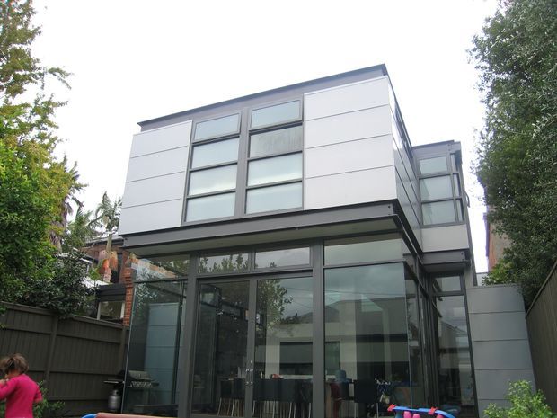 Exterior view of a contemporary multi-storey residence with a compact rectilinear form, flat roofline, large floor-to-ceiling glazed panels, and white horizontal panel cladding framed in dark grey. The house is surrounded by mature trees and enclosed fencing, with interior living and dining areas visible through the glazing.